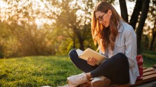 Woman with brown hair sitting next to a tree reading a book